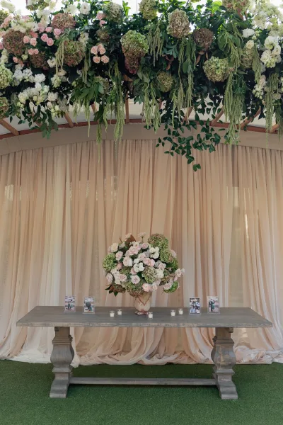 Wedding sweetheart table with a hanging floral installation over a rustic wood table, candles and framed photos beneath sheer tent draping