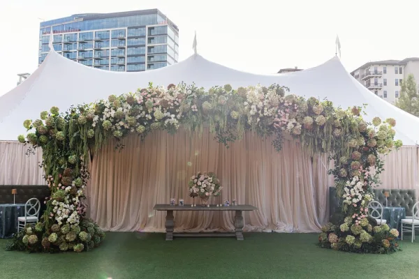 Wedding ceremony backdrop with floral arch and greenery, draped fabric, and candlelit sweetheart table under a white event tent