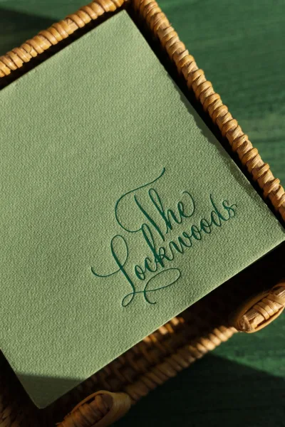Wedding invitation with green wedding invitation calligraphy on deckled edge paper, styled on a woven wicker tray over green fabric backdrop