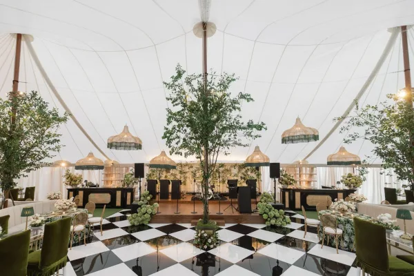 Reception decor in a wedding tent reception with hanging lanterns, candlelit banquet tables, greenery trees, and a checkered dance floor on lawn