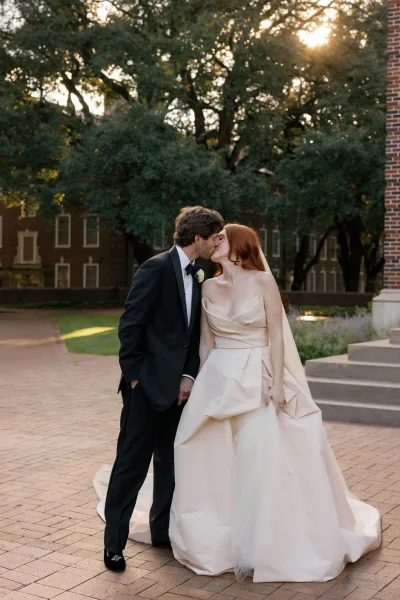 Wedding kiss portrait of bride and groom kissing, holding hands on sunlit brick courtyard steps, her long veil flowing behind