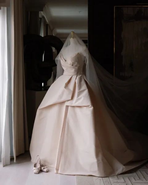 Wedding dress hanging on a hanger, a strapless ball gown with pleated satin skirt and long tulle veil in a hotel room by a window