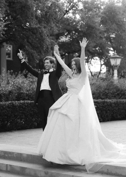 Wedding couple portrait in black and white with bride and groom raising hands, smiling in formal attire on stone steps in a garden setting
