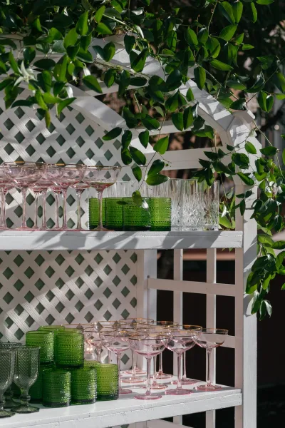 Wedding bar cart with champagne coupe glass display in pink and green glassware, set against a white lattice backdrop with greenery outdoors