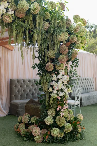 Wedding floral installation of hanging greenery and hydrangeas over a tufted velvet sofa, set beneath a draped outdoor tent in daylight