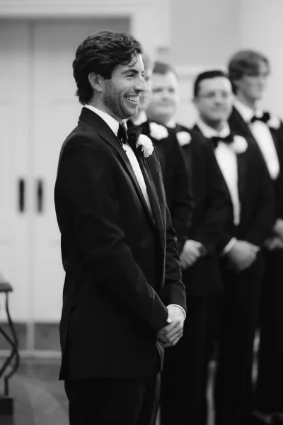 Groom portrait in a black and white wedding portrait, wearing a tuxedo with bow tie and white rose boutonniere beside groomsmen indoors