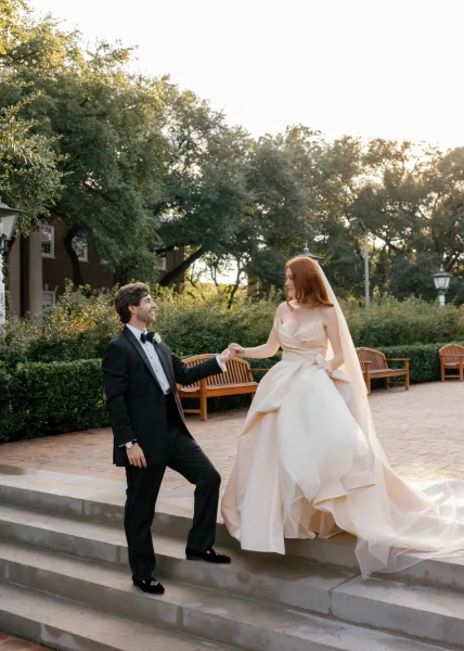 Couple portrait of bride and groom holding hands as he helps her up stone steps, long veil trailing in a garden brick courtyard