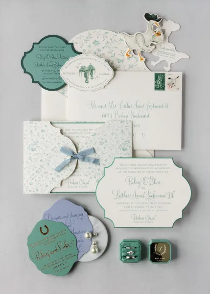 Wedding invitation suite with save the date card, envelopes, stamps, wax seal, ribbon bow, and rings arranged on a gray backdrop