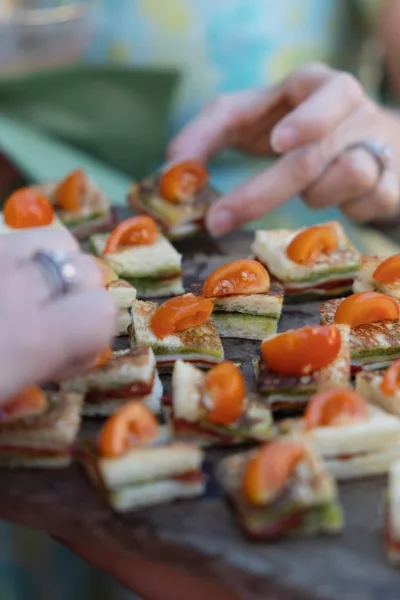 Wedding appetizers on a wooden serving board with finger sandwiches and cherry tomatoes, held by hands with rings at an outdoor cocktail hour