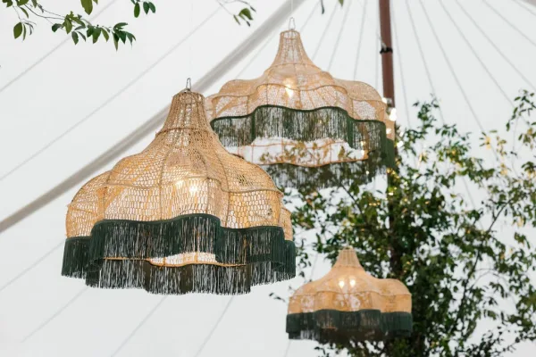 Hanging rattan lanterns and wicker pendant lights with fringe accents and string lights under a white tent canopy, trees beyond