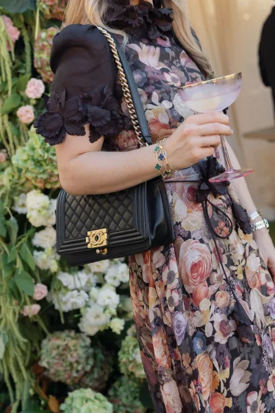 Wedding guest outfit in a floral wedding guest dress with black 3D appliques, holding a cocktail glass by a flower wall backdrop with guests