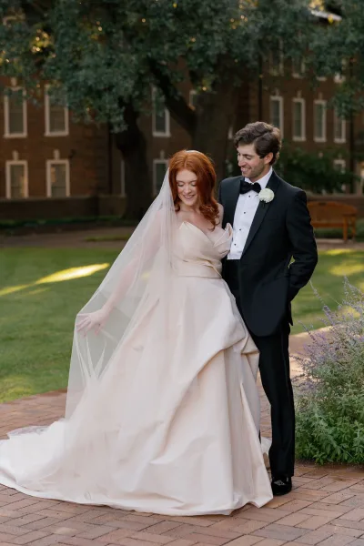 Couple portrait of bride in a satin gown holding her veil as groom in tuxedo looks at her on a brick walkway in a garden courtyard