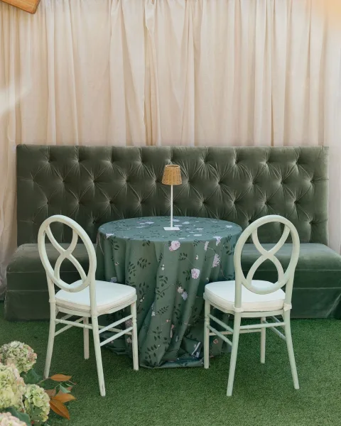 Wedding lounge seating with tufted velvet banquette, round table in floral cloth, white chairs and lamp before cream draping on green turf.