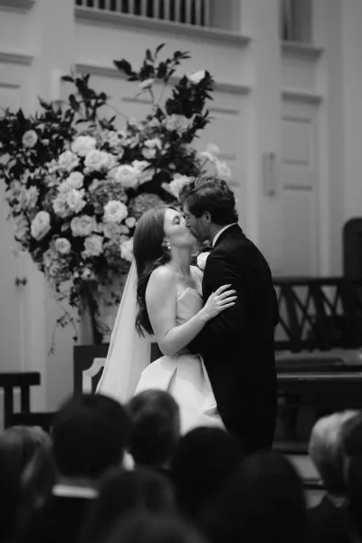 Wedding kiss as bride in veil and strapless gown embraces groom in tuxedo, framed by large floral arrangement and seated guests indoors