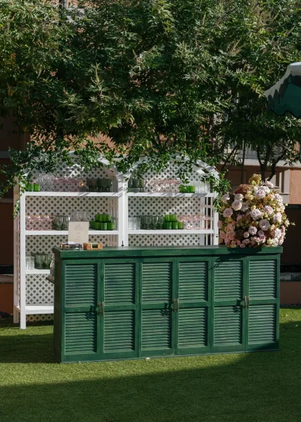 Wedding bar setup with emerald green bar counter, glassware on white lattice shelves, green votive candles and floral menu card outdoors