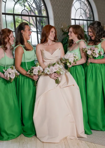 Bride with bridesmaids in a bridal party portrait, laughing in emerald green satin dresses with bouquets in a room with arched windows