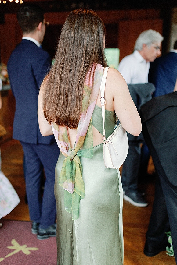 Wedding guest outfit featuring a sage green wedding guest dress in satin slip style, with pastel scarf, white shoulder bag, and bistro-lit reception backdrop