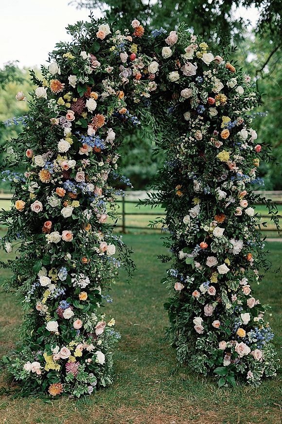 Wedding floral arch with lush greenery, roses, wildflowers and baby’s breath on a grassy lawn before a wooden fence and trees