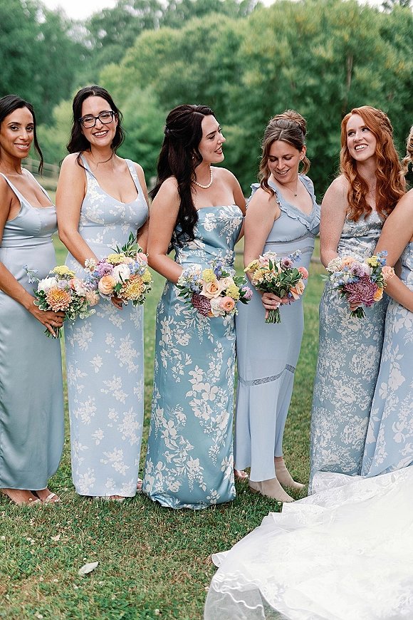 Bridesmaid group photo with dusty blue bridesmaid dresses, holding colorful bouquets on a grassy lawn with trees behind