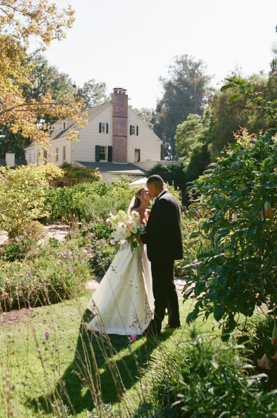 Wedding kiss portrait of newlyweds on a garden pathway, bride holding bouquet as trees frame the house exterior in soft natural light.