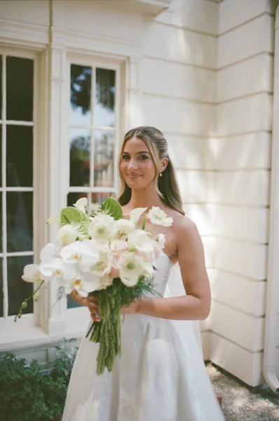 Bridal portrait of a bride holding a bouquet with white orchids, standing by a house window with white siding and garden plants along a gravel path.