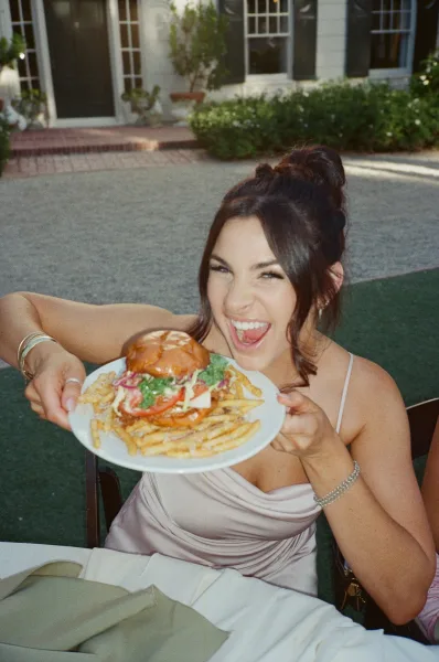 Reception candid of a laughing wedding guest with a burger, in a garden courtyard with house facade and shrubs at an outdoor table.