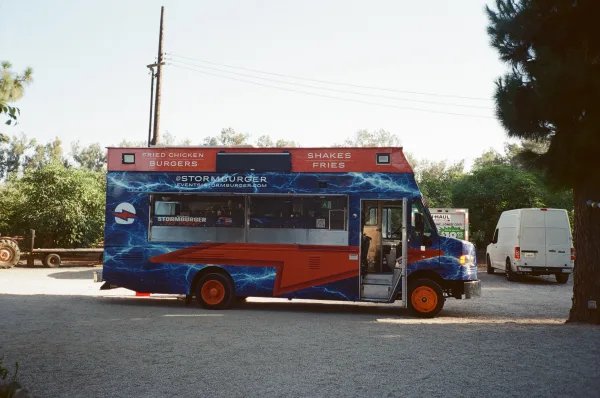 Wedding food truck at a casual food truck reception, open service window, parked on a gravel lot with trees and vans under a clear sky.
