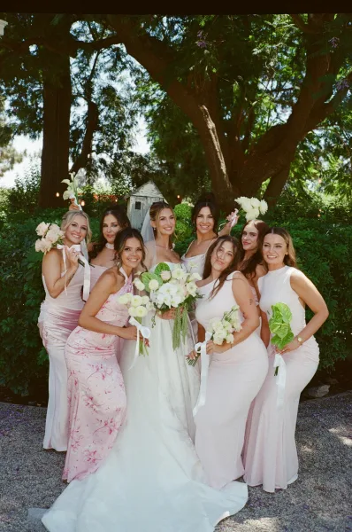 Bridal party portrait with light pink bridesmaid dresses surrounds a bride with a veil on a gravel path between garden hedges and trees.