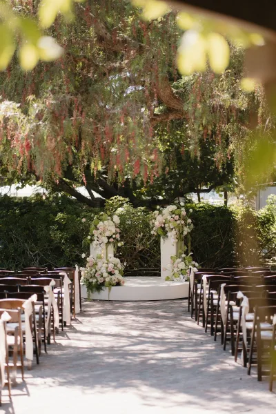 Ceremony setup for an outdoor garden ceremony on a round platform, framed by garden trees and hedges, with hanging branches over stone pavers.