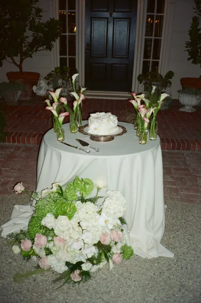 Wedding cake table with a small wedding cake on a silver cake stand, set on a brick patio by a house front door and potted plants