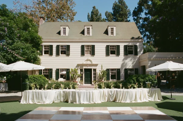 Outdoor reception setup centers a reception with dance floor and market umbrellas before an estate house facade and garden shrubs.