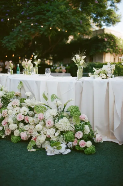 Reception table decor for an outdoor reception tablescape featuring bud vases beneath garden trees and warm string lights.