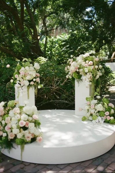Ceremony setup with wedding altar flowers flanking a round platform, set on a brick patio amid garden trees in soft, dappled sunlight.
