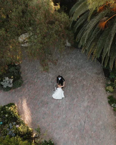 First dance on a brick courtyard as the couple twirls, a sweeping bridal gown flowing amid garden greenery beneath a palm tree and leafy canopy.