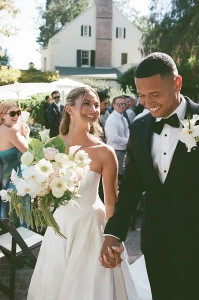 Wedding recessional with bride and groom walking hand in hand past guests' parasols on a brick patio amid garden greenery, beaming with joy.
