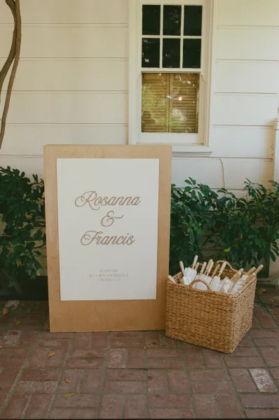 Wedding welcome sign in neutral wedding signage style, accompanied by paper parasols on a brick patio against a white siding wall and window.