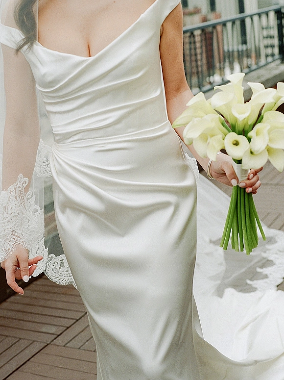Wedding dress in silk satin with a cowl neck, shown with lace veil and calla lily bouquet on an outdoor balcony by metal railing