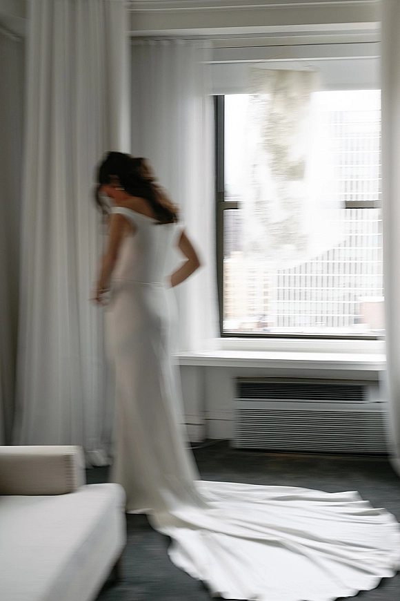 Wedding dress, lace wedding dress with off-the-shoulder sleeves and long train by a large hotel window with sheer curtains and city view