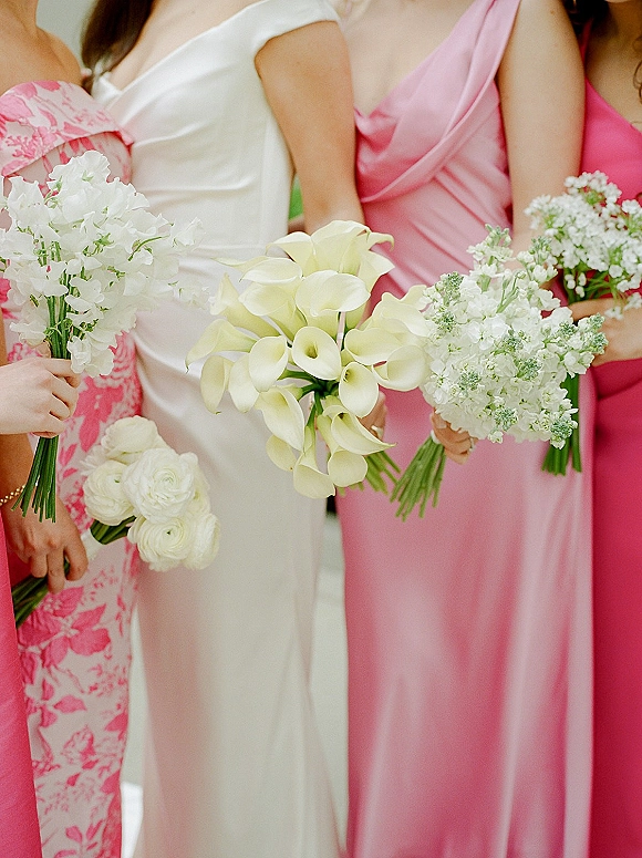 Bridesmaid bouquets of white calla lily bouquet and ranunculus held by women in pink dresses, with blurred garden greenery behind