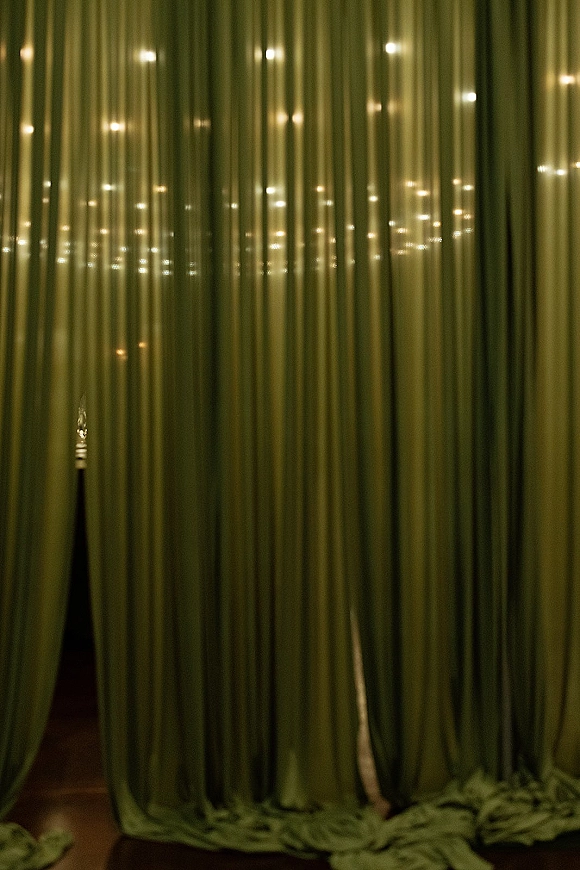 Wedding drapery backdrop with green velvet drape backdrop, ceiling-to-floor fabric and warm string lights glowing in a dark room