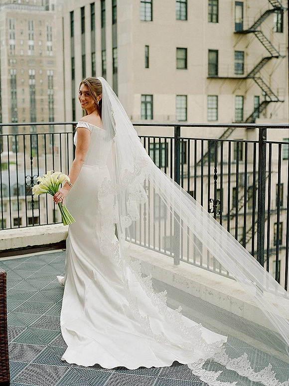 Bridal portrait of a bride in an off shoulder wedding dress with a long lace veil, holding a calla lily bouquet on a city rooftop terrace balcony