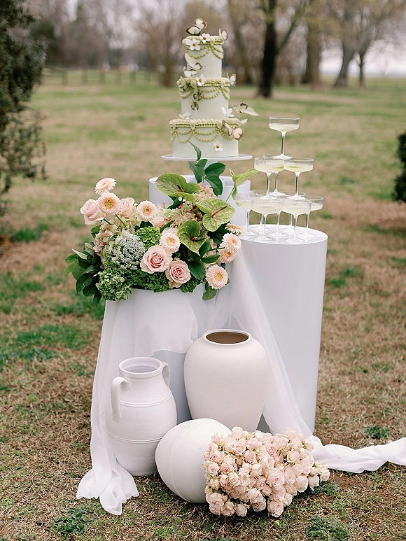 Wedding cake display with a tiered wedding cake on a stand beside a champagne coupe tower, roses and greenery on a grassy lawn