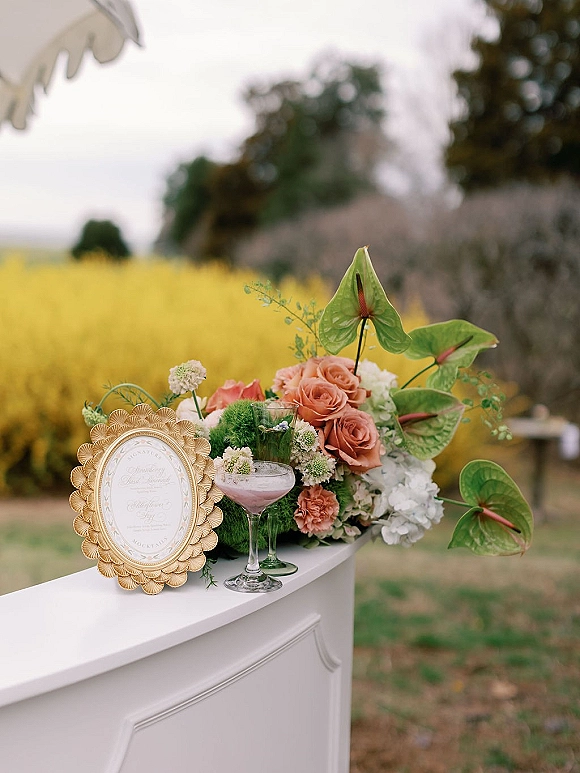 Cocktail bar decor with a framed menu in a gold ornate frame, bar-top florals, and coupe and highball cocktails on an outdoor lawn