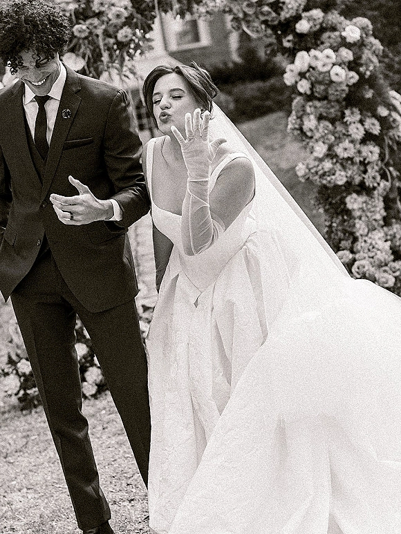 Couple portrait of bride showing wedding ring as she blows a kiss, long veil and gloves beside groom under a floral arch in garden