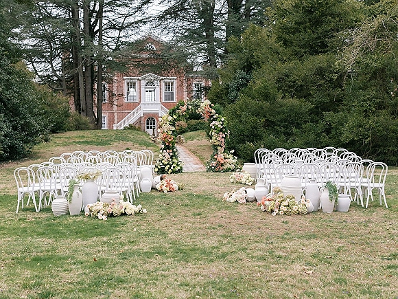 Ceremony setup with outdoor wedding ceremony setup featuring a floral arch, white chairs, and aisle runner on a garden lawn by a brick house