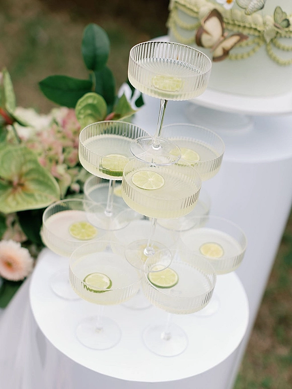 Champagne tower setup with stacked coupe glass tower, lime-garnished cocktails, and wedding cake on a white pedestal outdoors on grass lawn