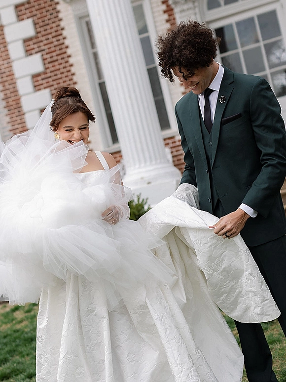 Couple portrait of bride and groom laughing as he lifts her tulle skirt, her veil flowing, on steps by a brick building with columns