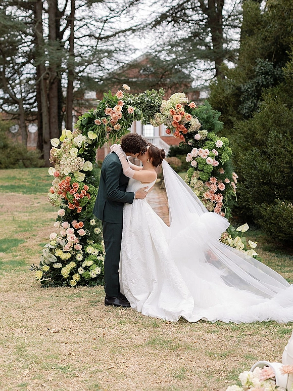 Wedding kiss portrait of bride and groom kissing under a rose and hydrangea floral arch, cathedral veil trailing on a garden lawn by a brick building