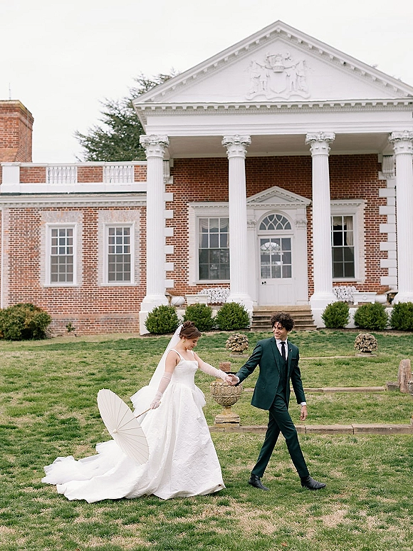 Couple portrait of bride and groom walking hand in hand, bride holding a parasol and veil flowing before a brick mansion with columns