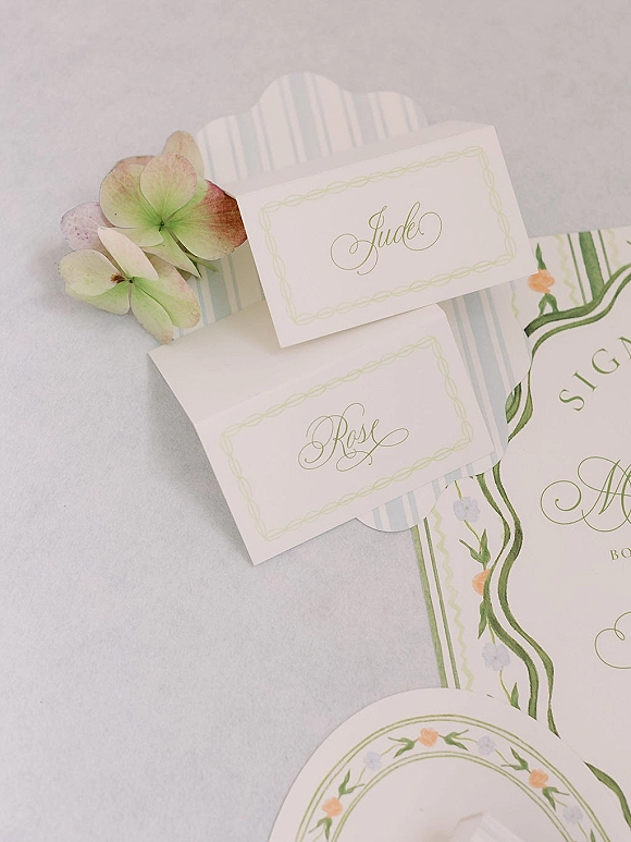 Wedding place cards with calligraphy place cards beside a floral invitation and monogram crest on a white linen tablecloth, styled with hydrangea blooms